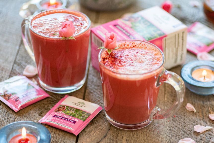 Two mugs of pink tea with rosebuds on a wooden table, accompanied by tea packets and lit candles.