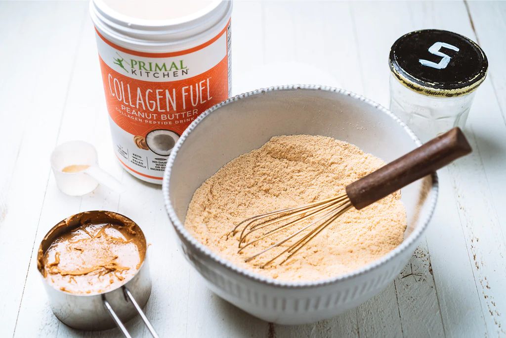 Ingredients for a recipe: a container of collagen fuel, bowl with whisked mixture, a measuring cup, and salt shaker on a white table.