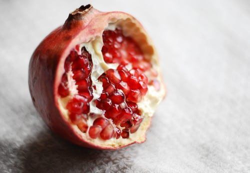 Half-eaten pomegranate with bright red seeds and a thick white membrane, set on a textured light background.