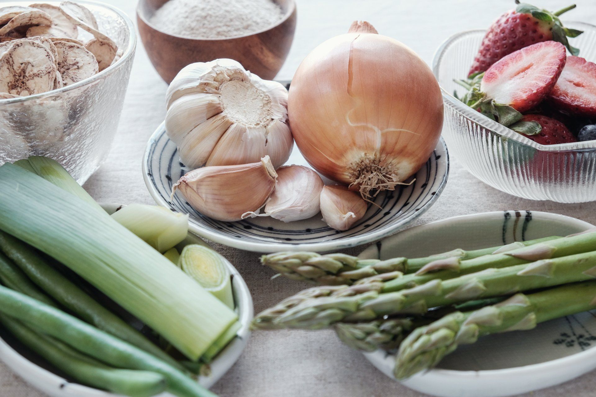 Close-up of fresh produce: garlic, onion, asparagus, leeks, strawberries, chips, and flour in small bowls.