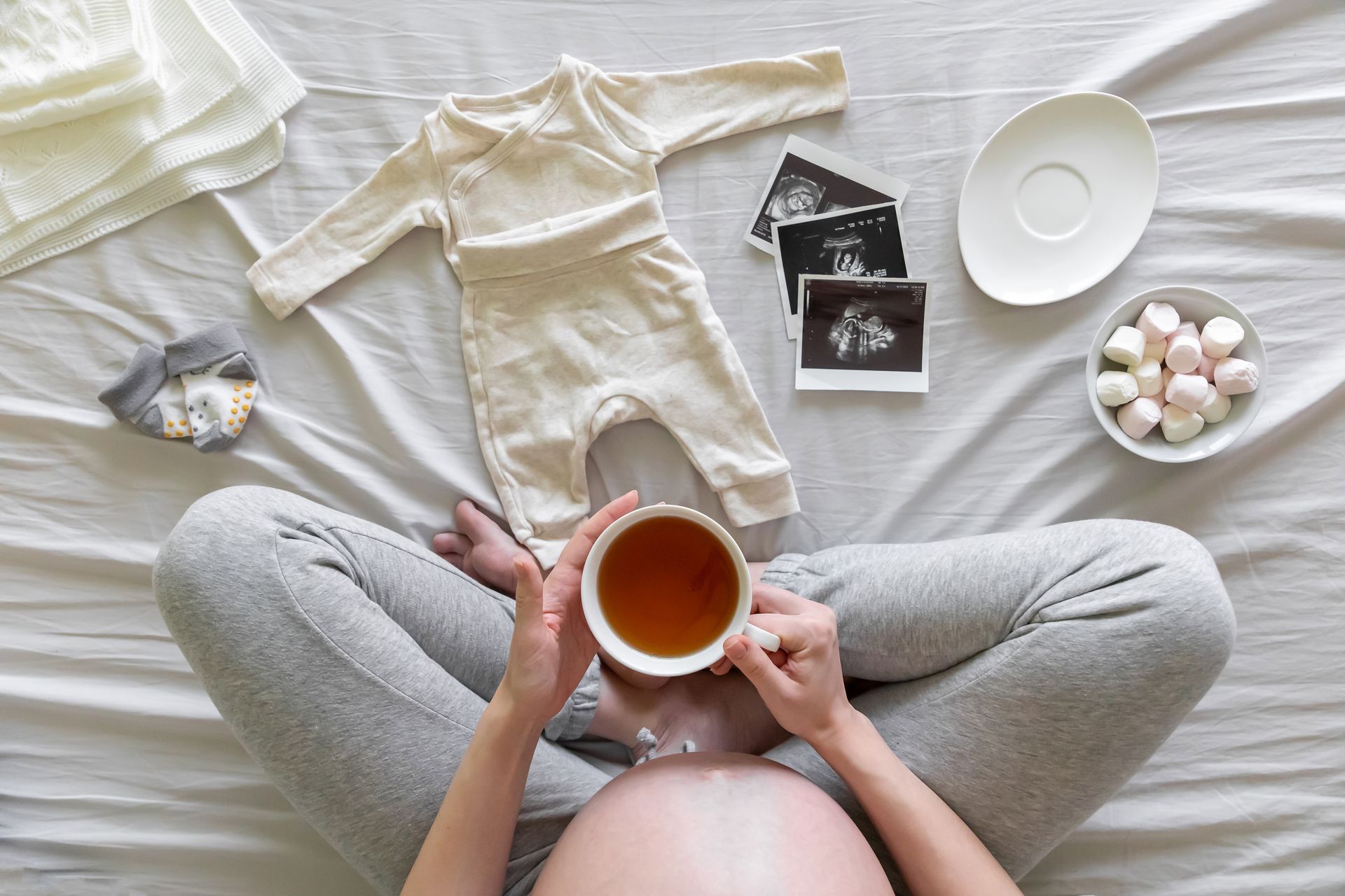 Pregnant person holding tea, surrounded by baby clothes, ultrasound photos, and snacks, on a bed.