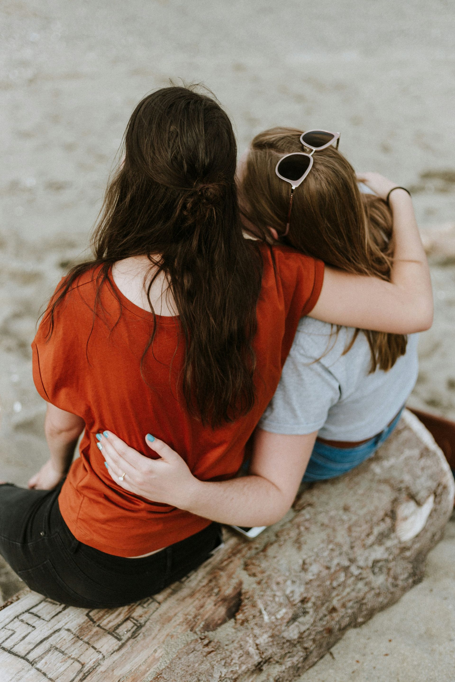 Two women with arms around each other sit on a log at the beach. One wears red, the other gray, both facing the water.