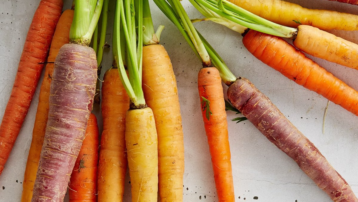 Close-up of fresh, colorful carrots: orange, yellow, and purple with green tops, scattered on a light surface.