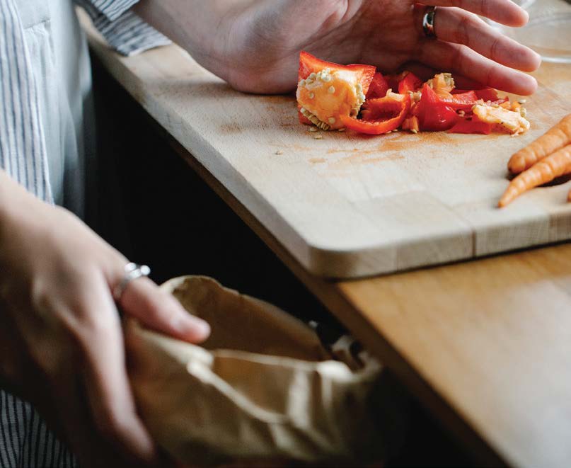 Hands collecting food scraps from a cutting board, a red pepper being chopped and carrots visible.