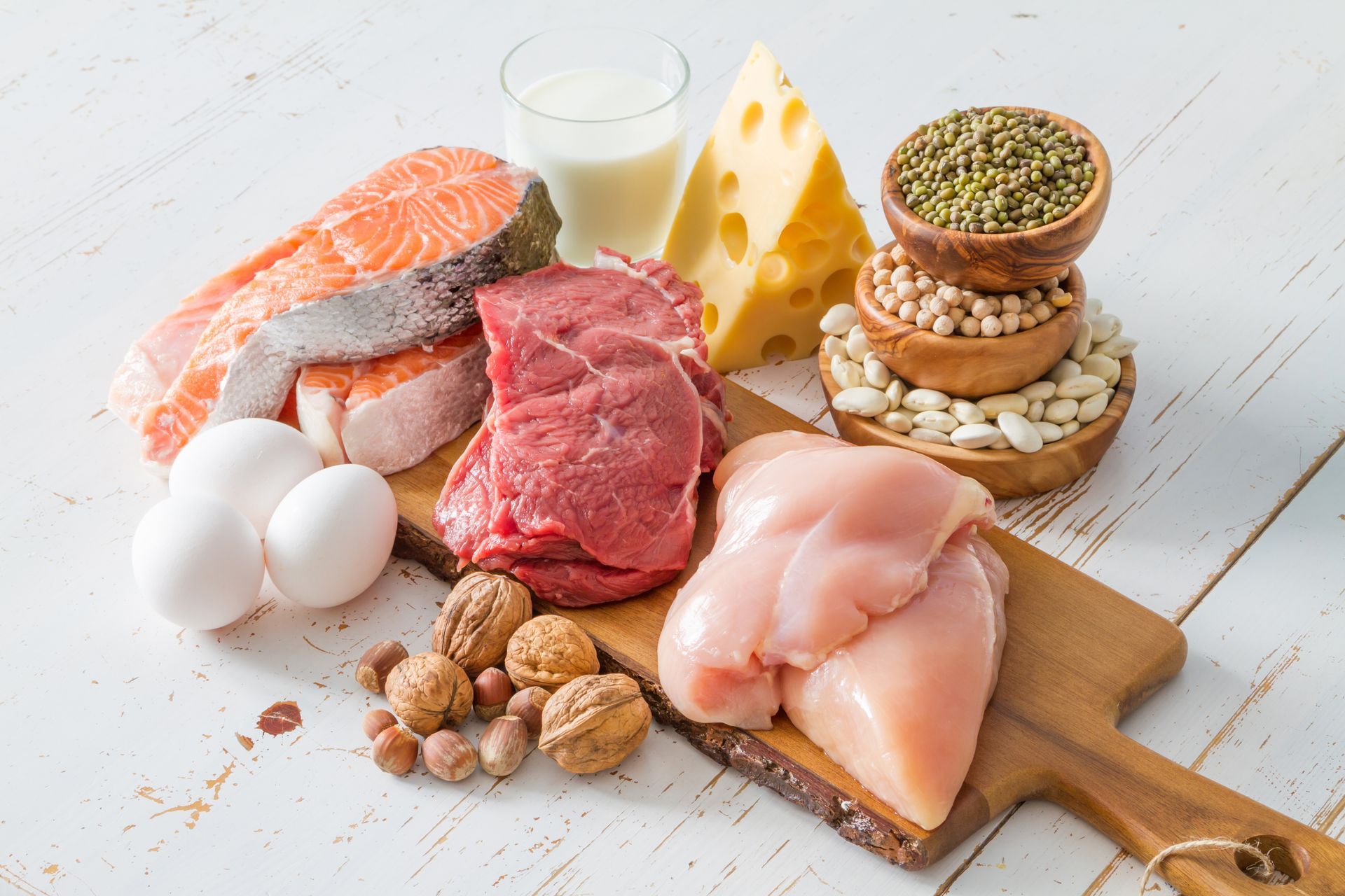 Assortment of protein-rich foods: salmon, steak, chicken breast, eggs, cheese, beans, nuts, and milk.
