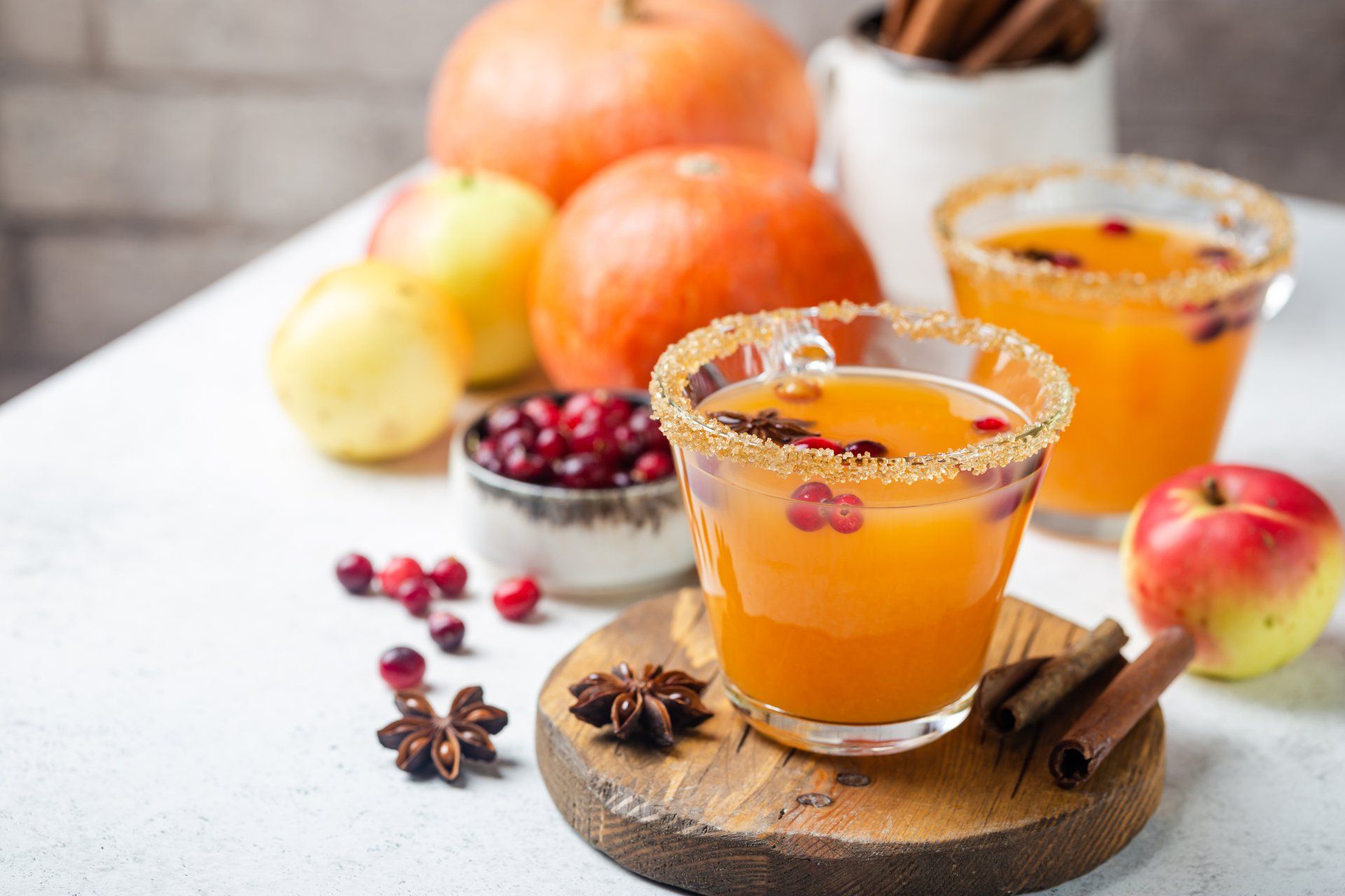 Two glasses of pumpkin spiced drink with sugared rims, garnished with cranberries. Pumpkins, apples, cinnamon sticks and star anise surround the drinks.