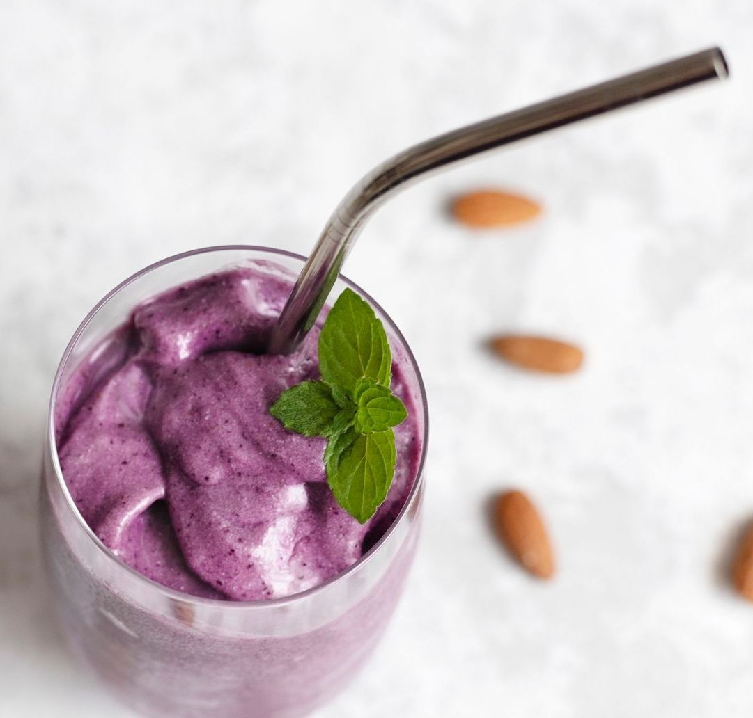 Purple smoothie in a glass with a metal straw and mint sprig, with almonds scattered nearby.