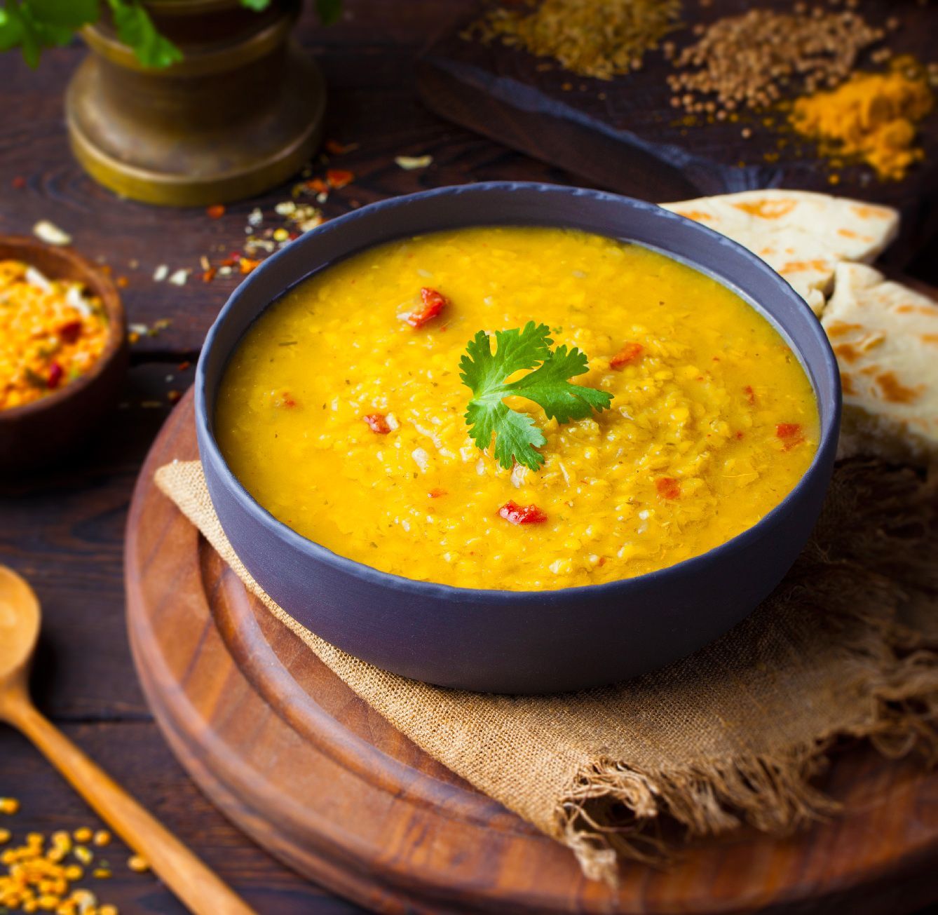 A bowl of yellow lentil soup garnished with cilantro, served on a wooden board with naan and spices.