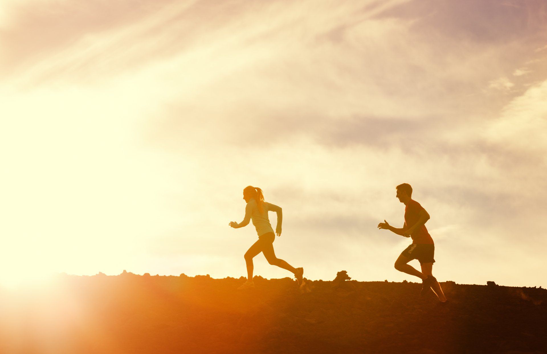 Two runners silhouetted against a bright sky, jogging uphill. Golden sunlight illuminates the scene.
