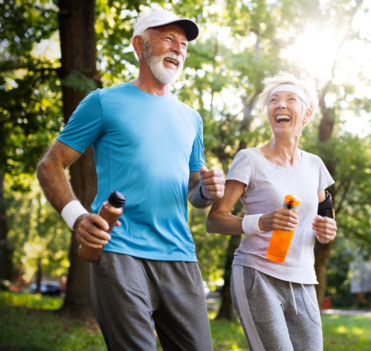An elderly couple in athletic wear running and laughing in a park on a sunny day.