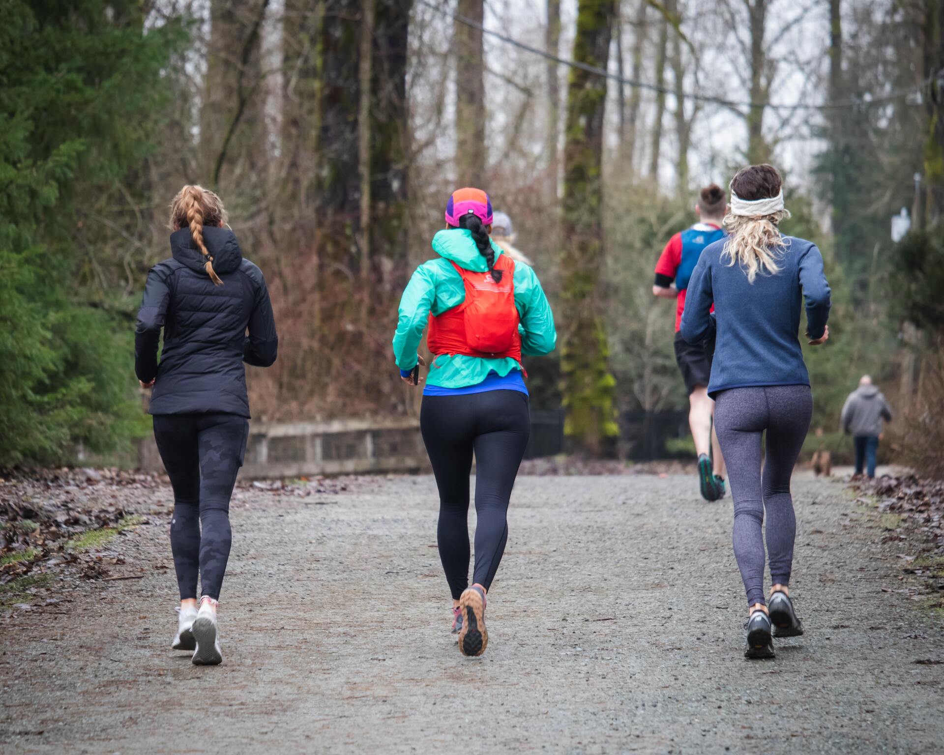 People run on a gravel path through a forest. Runners wear activewear, including leggings and jackets.