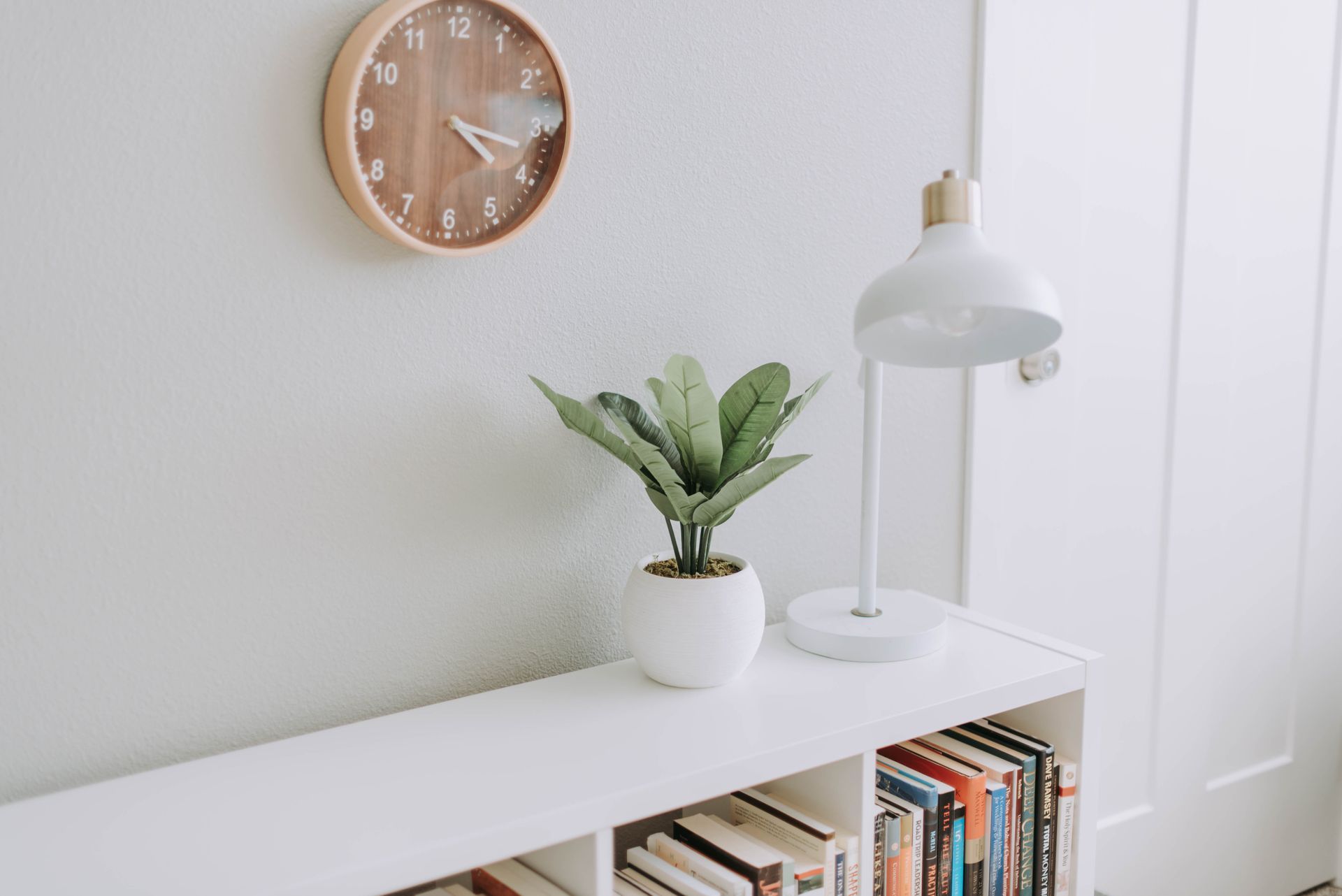 A white shelf with a plant, lamp, and books. A wooden clock hangs on the light gray wall.