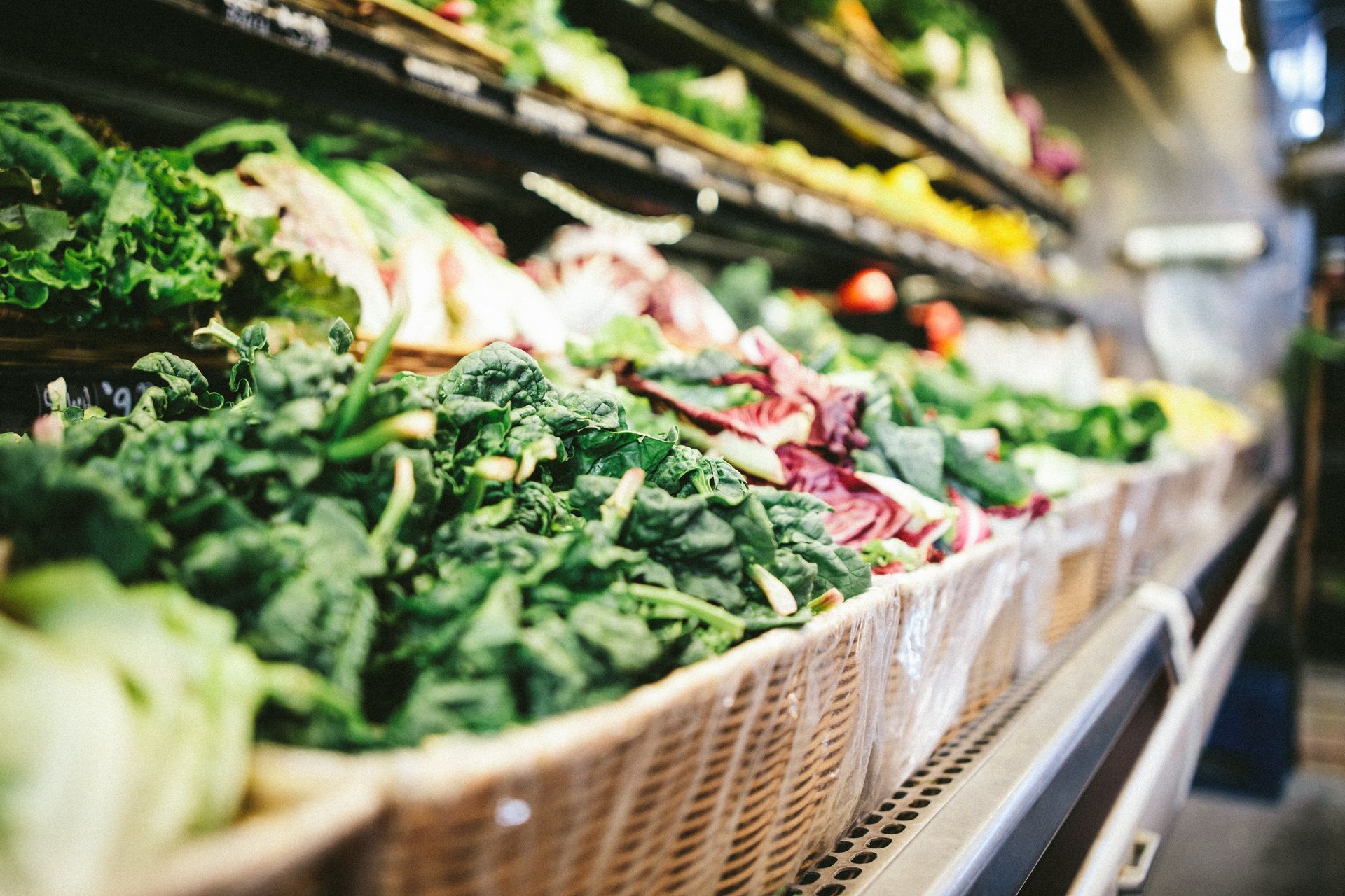 Fresh produce, including spinach, lettuce, and other greens, is displayed in a supermarket produce section. The scene is brightly lit with baskets filled with vegetables.