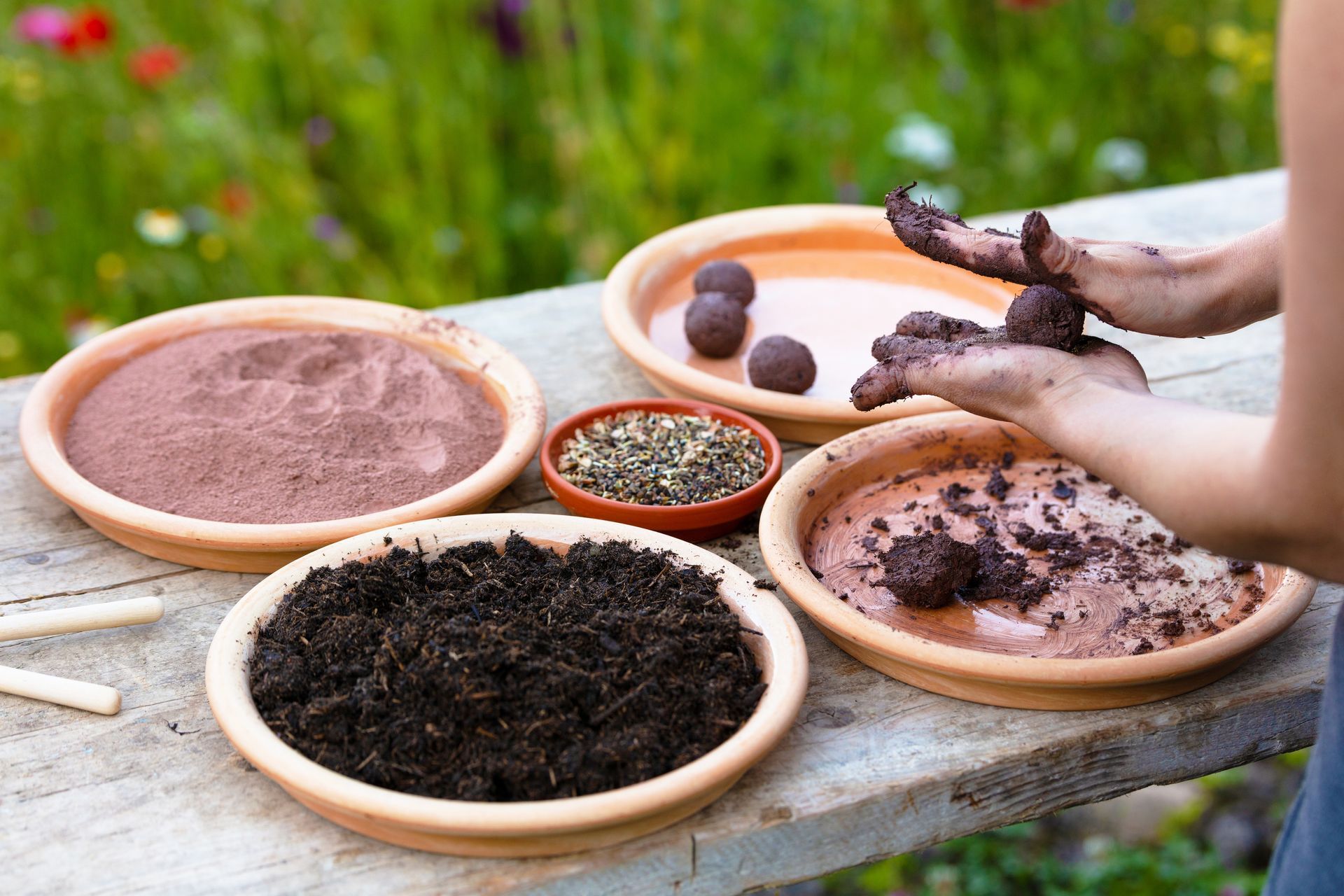 Hands rolling seed balls, surrounded by bowls of soil, powder, and seeds on a wooden table outdoors.