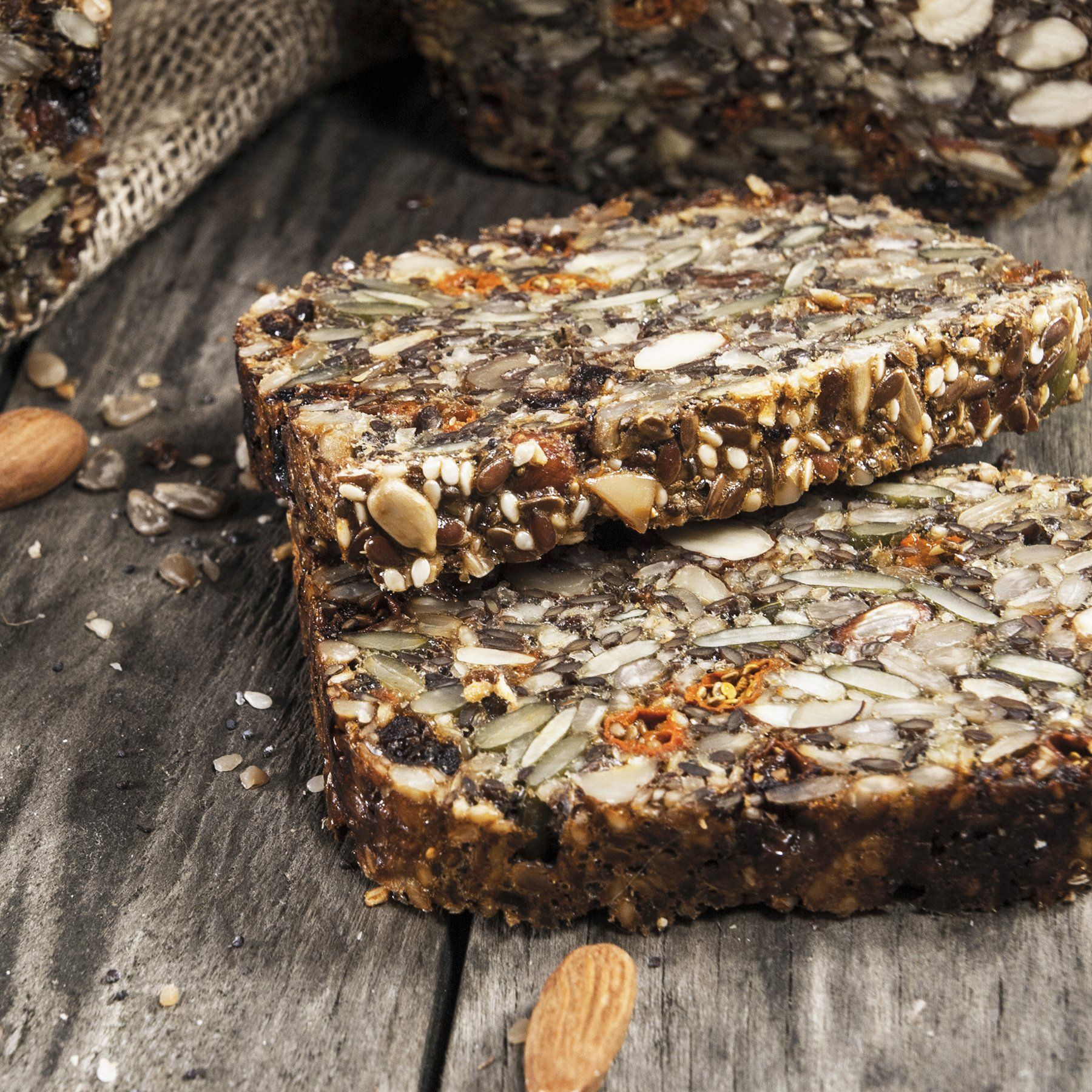 Two slices of seed and nut bread stacked on a weathered wooden surface, with scattered almonds and other bread pieces nearby.