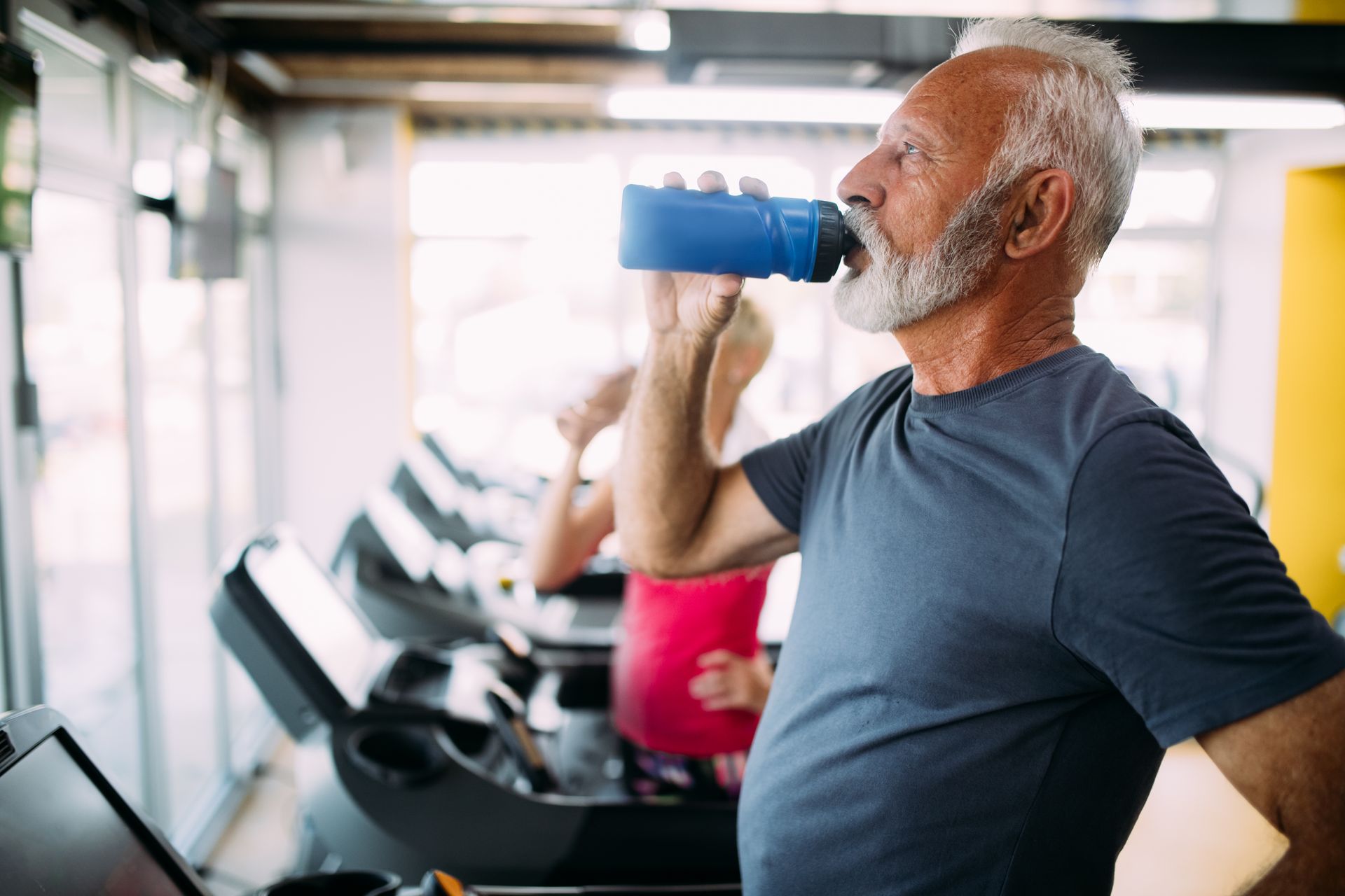 Older man in a gym drinks from a blue water bottle while standing near a treadmill, another person runs in the background.