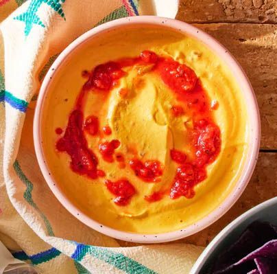 A bowl of creamy yellow dip with red berry sauce. It sits on a colorful patterned cloth, with a bowl of purple chips nearby.