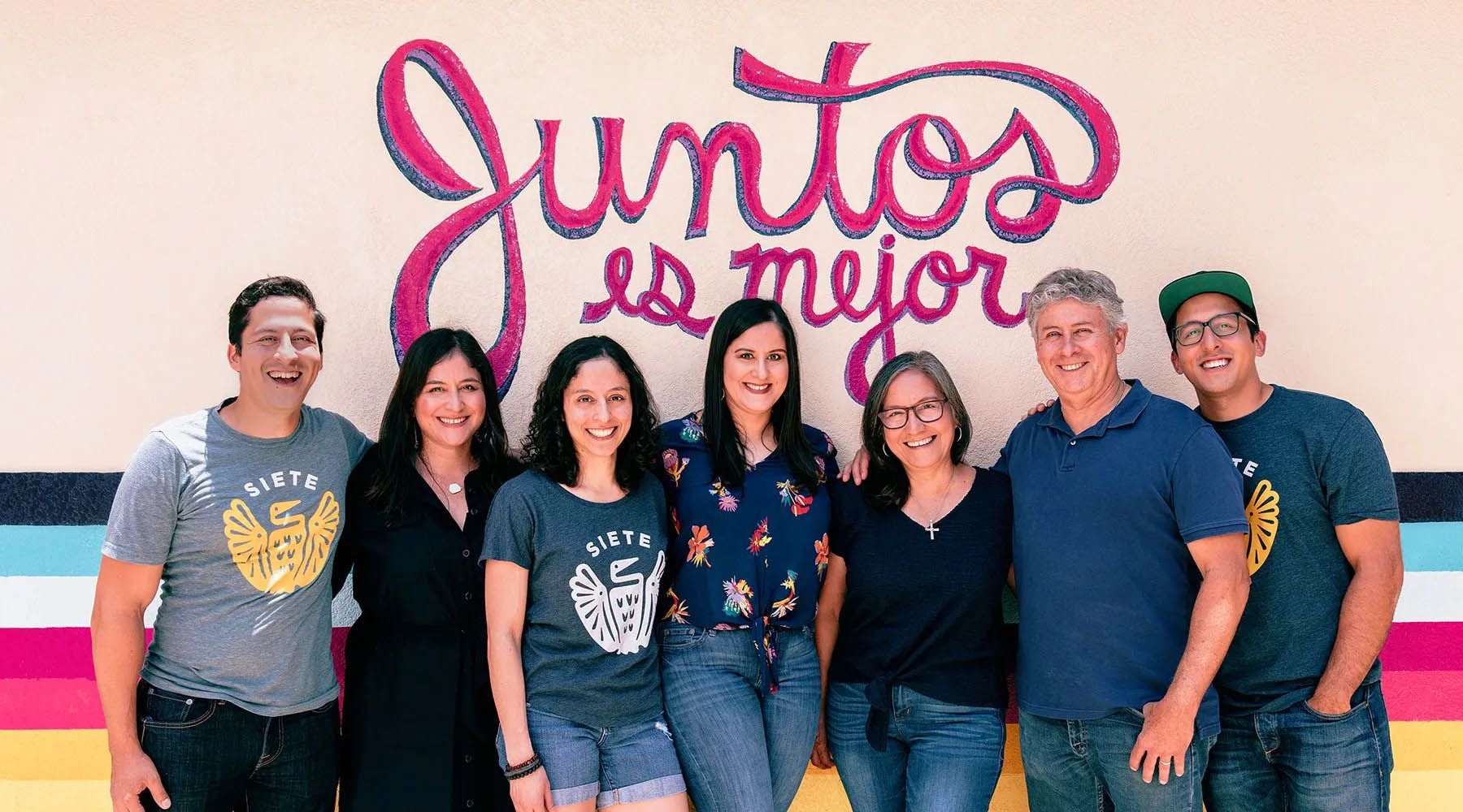 Group of seven people smiling and posing in front of a colorful mural that reads 