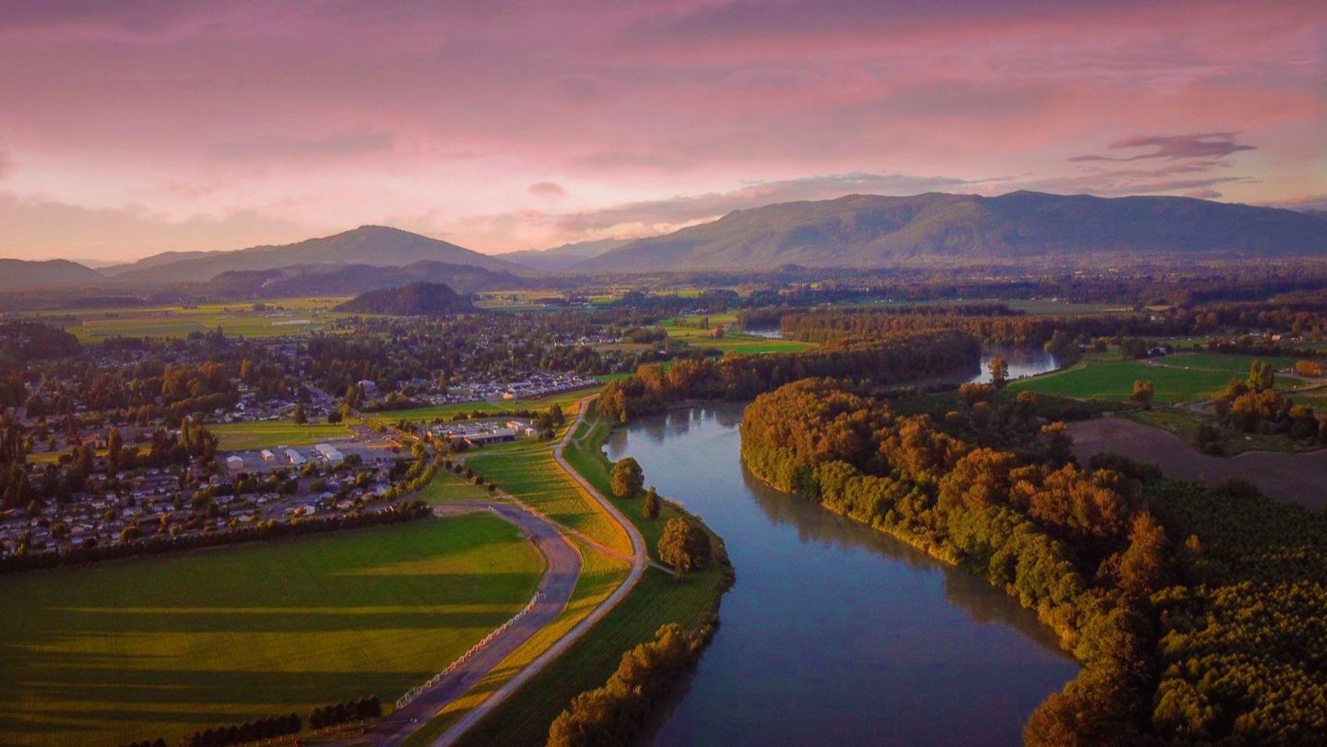 River flows through a valley with a town on the left and trees on the right, under a colorful sunset.