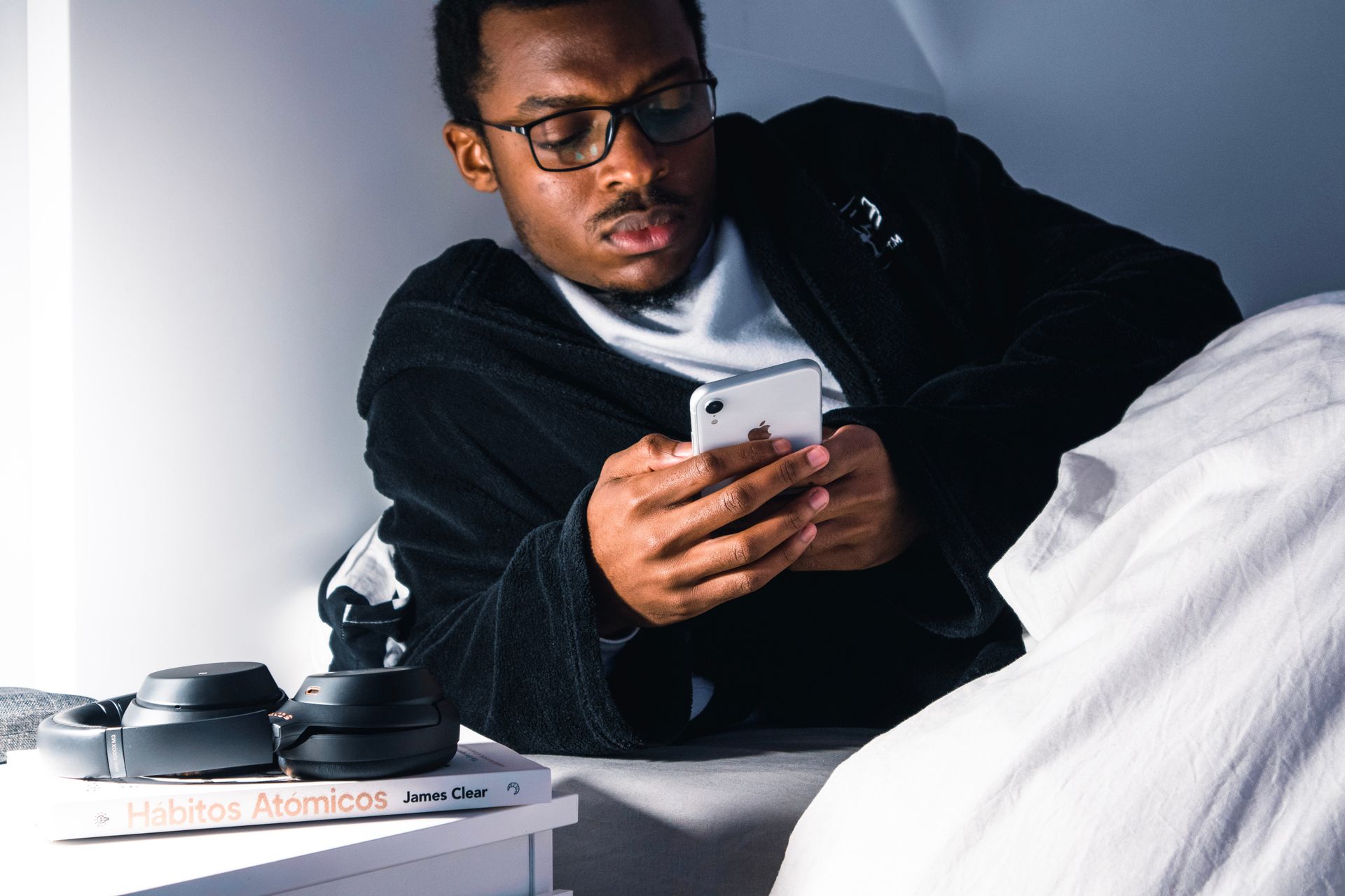 A Black man in glasses and a robe looks at his phone while lying in bed. Headphones and a book sit on the bedside table.