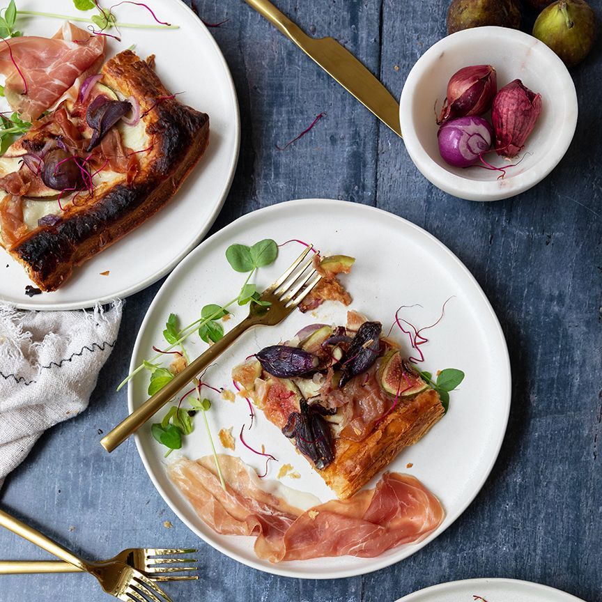 A close-up of a fig and prosciutto tart on a white plate, garnished with microgreens and served with a golden fork.