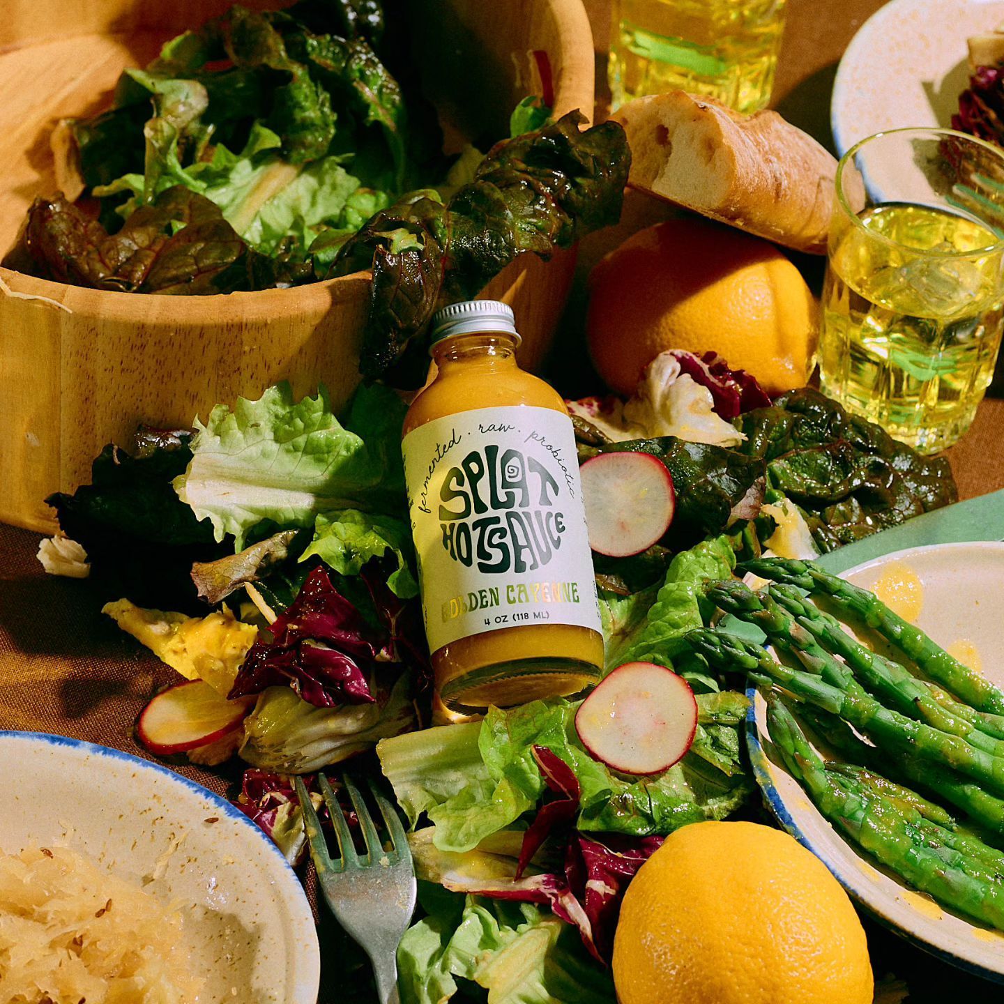 A close-up of a vibrant salad with asparagus, radishes, greens, and a bottle of dressing, alongside bread, lemons, and drinks.