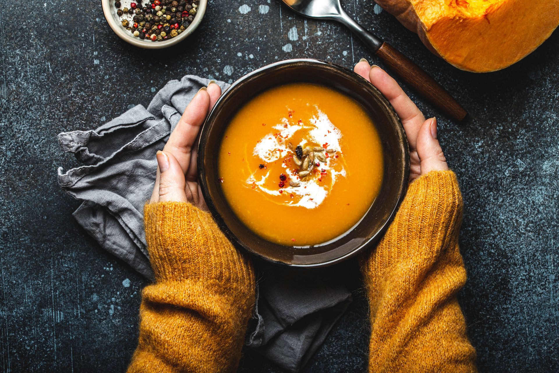 Hands holding a bowl of orange soup, likely pumpkin, on a dark surface, next to spices and a pumpkin slice.