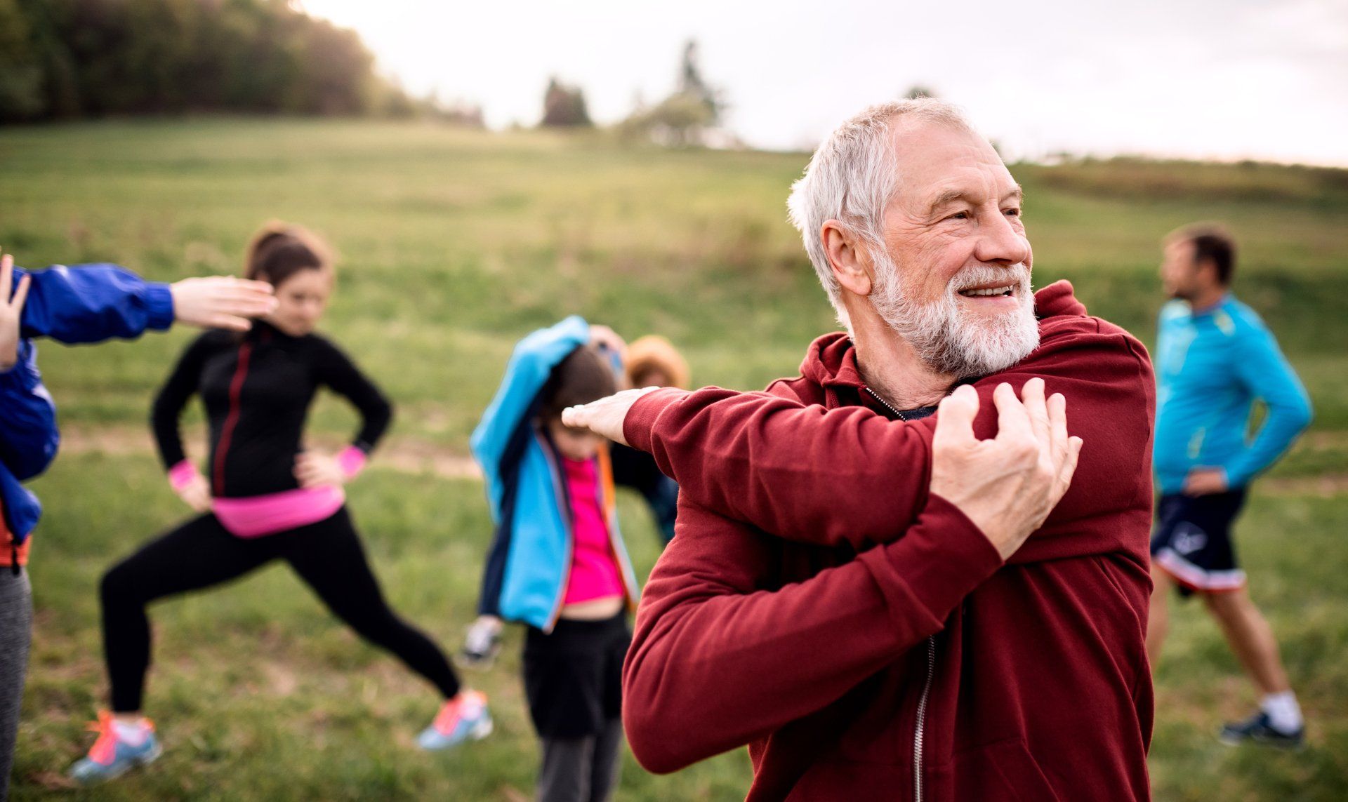 An older man with a gray beard smiles while stretching outdoors with a group of people. He wears a red hoodie.