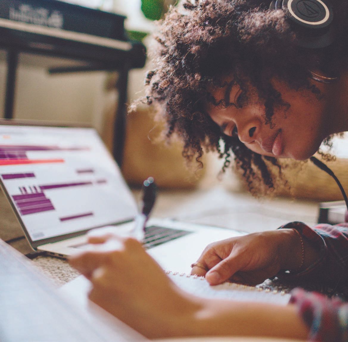 Person with headphones and curly hair, writing in a notebook while looking at a laptop screen.