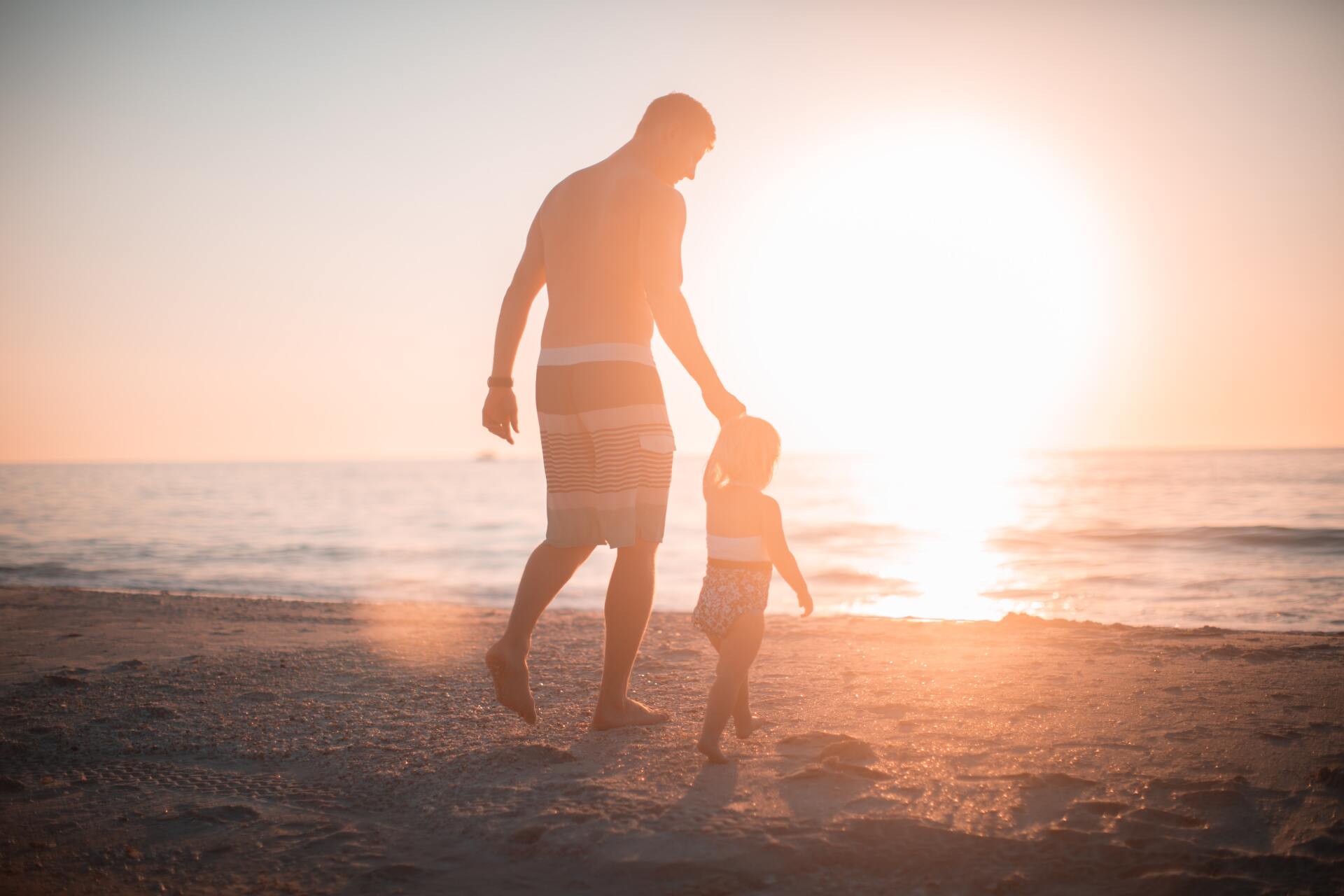 Father and child walking on a beach at sunset, silhouetted against the bright sun.