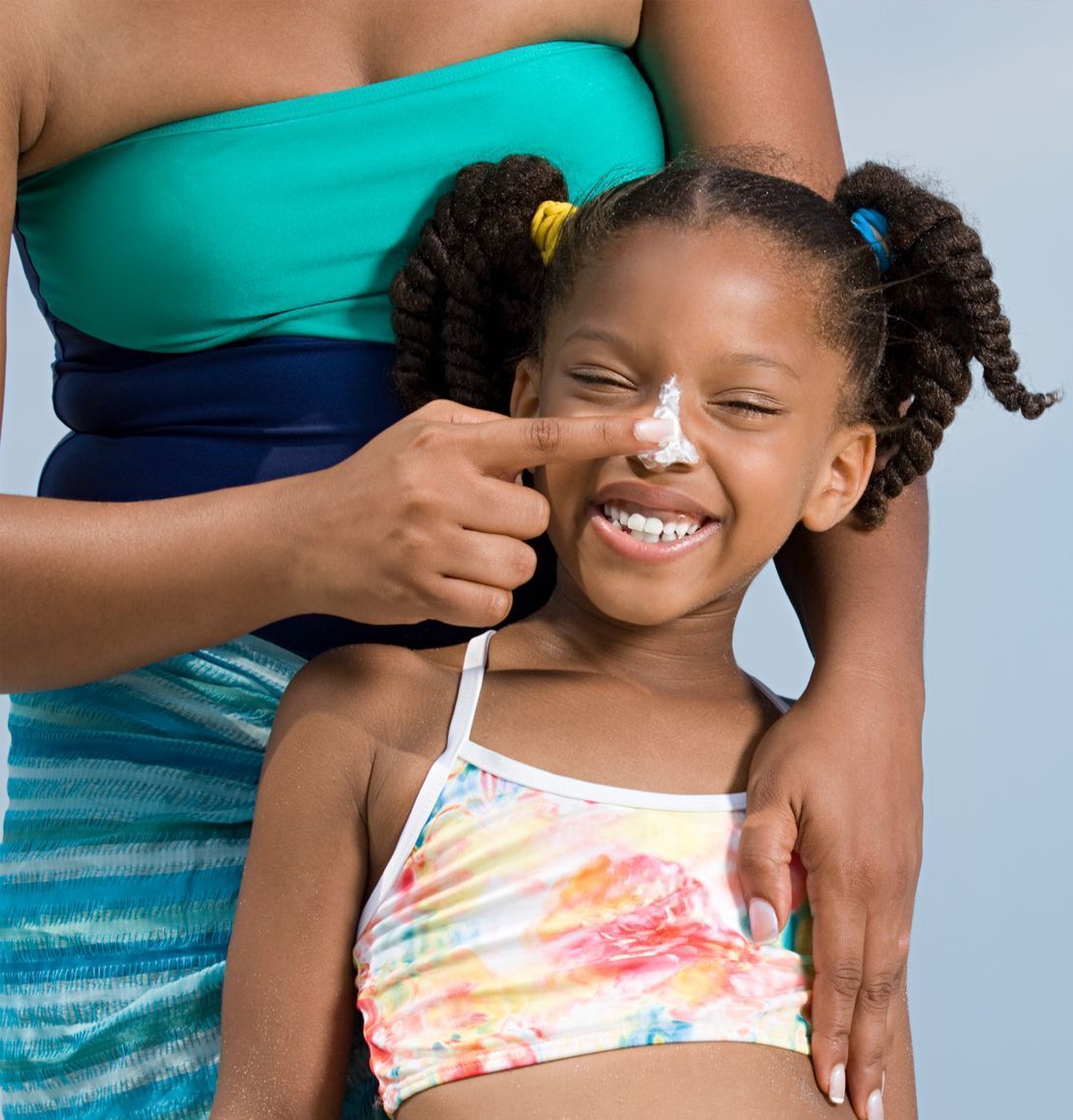 Woman applying sunscreen to a smiling girl's nose. Both are outdoors; the girl wears a swimsuit.