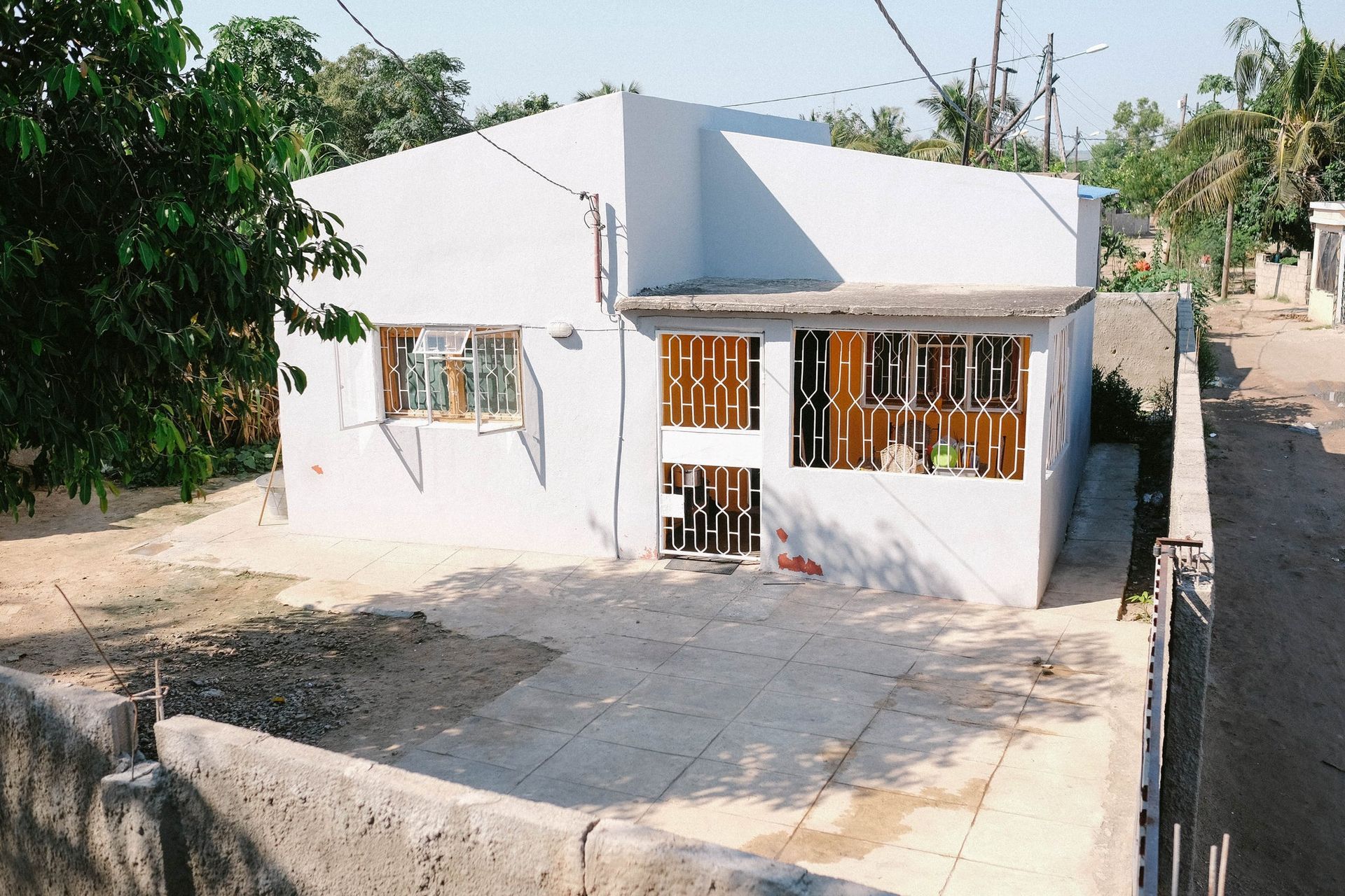A small, whitewashed house with a concrete patio and partial fence in a sunny outdoor setting.