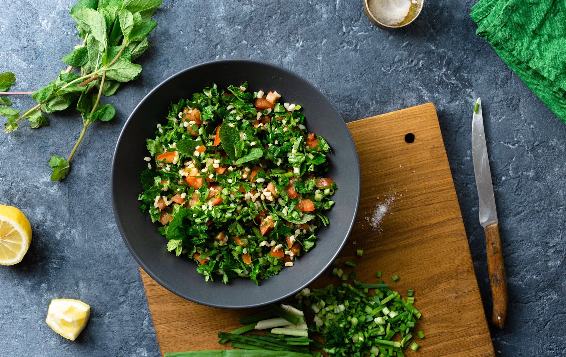 Bowl of tabbouleh salad on a wooden cutting board with fresh mint, tomatoes, and lemon on a dark gray surface.