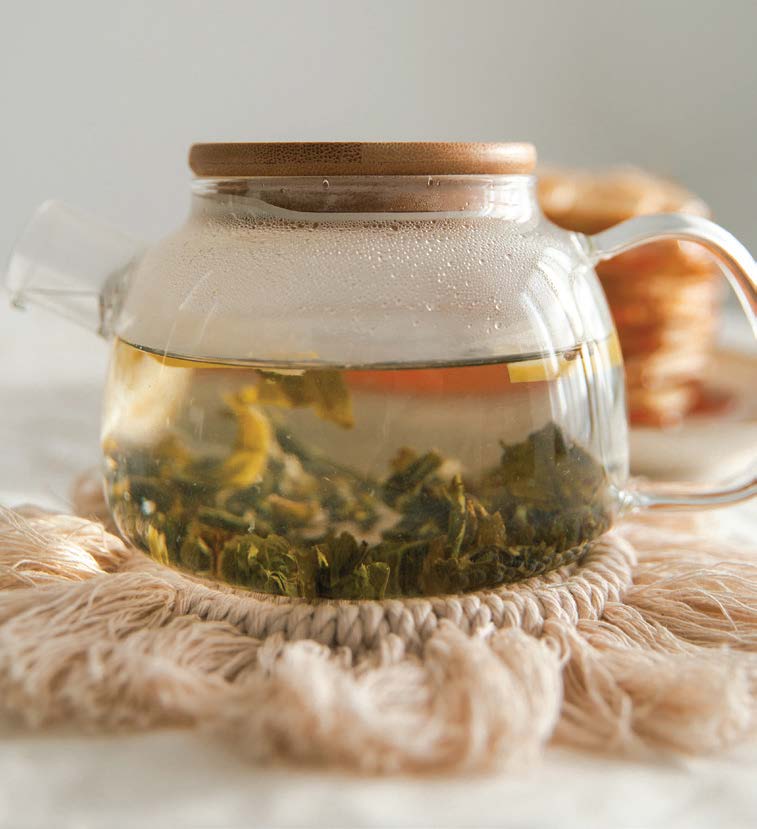 A glass teapot brewing green tea on a beige macrame coaster, with a stack of cookies in the background.