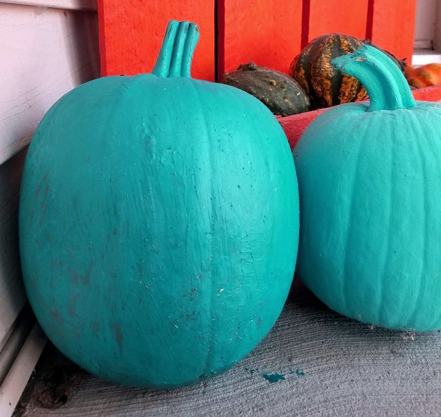 Two teal painted pumpkins on a porch with an orange background and other gourds.