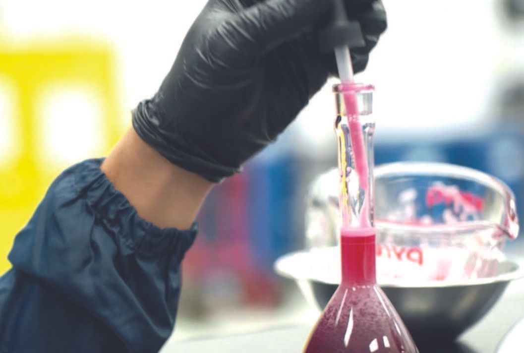 Person in black gloves uses a dropper to add pink liquid to a flask containing a darker liquid. Laboratory setting.