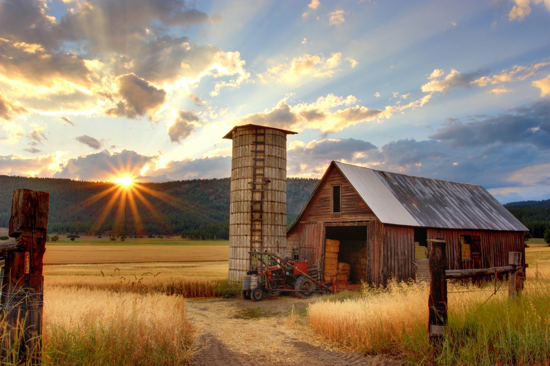 Sunset over a rustic barn and silo in a golden field, with a wooden fence in the foreground.