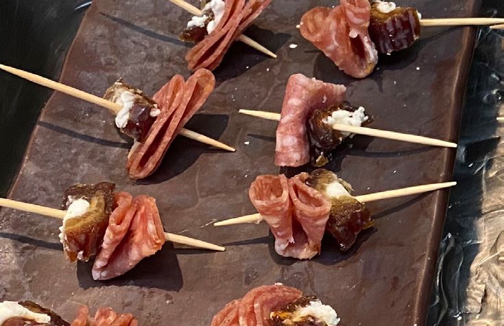 Skewered appetizers: dates filled with cheese, wrapped in salami, on a dark brown serving board.