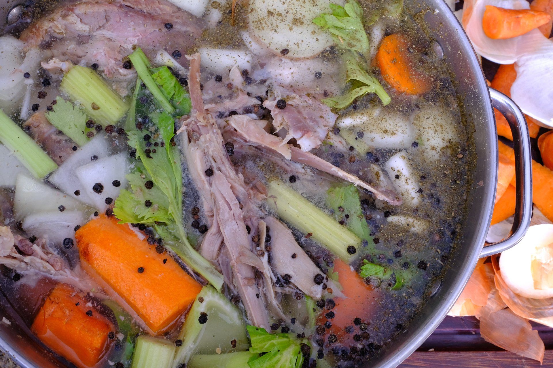 Close-up of a metal pot filled with chicken broth, vegetables, and shredded chicken. Carrots, celery, onion, and peppercorns are visible.