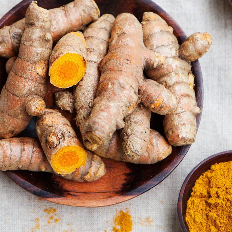 Fresh turmeric root in a wooden bowl, with ground turmeric in a small bowl at the bottom right.