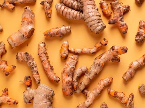 Fresh turmeric roots scattered on an orange surface, showing their knobby shape and orange-yellow flesh.
