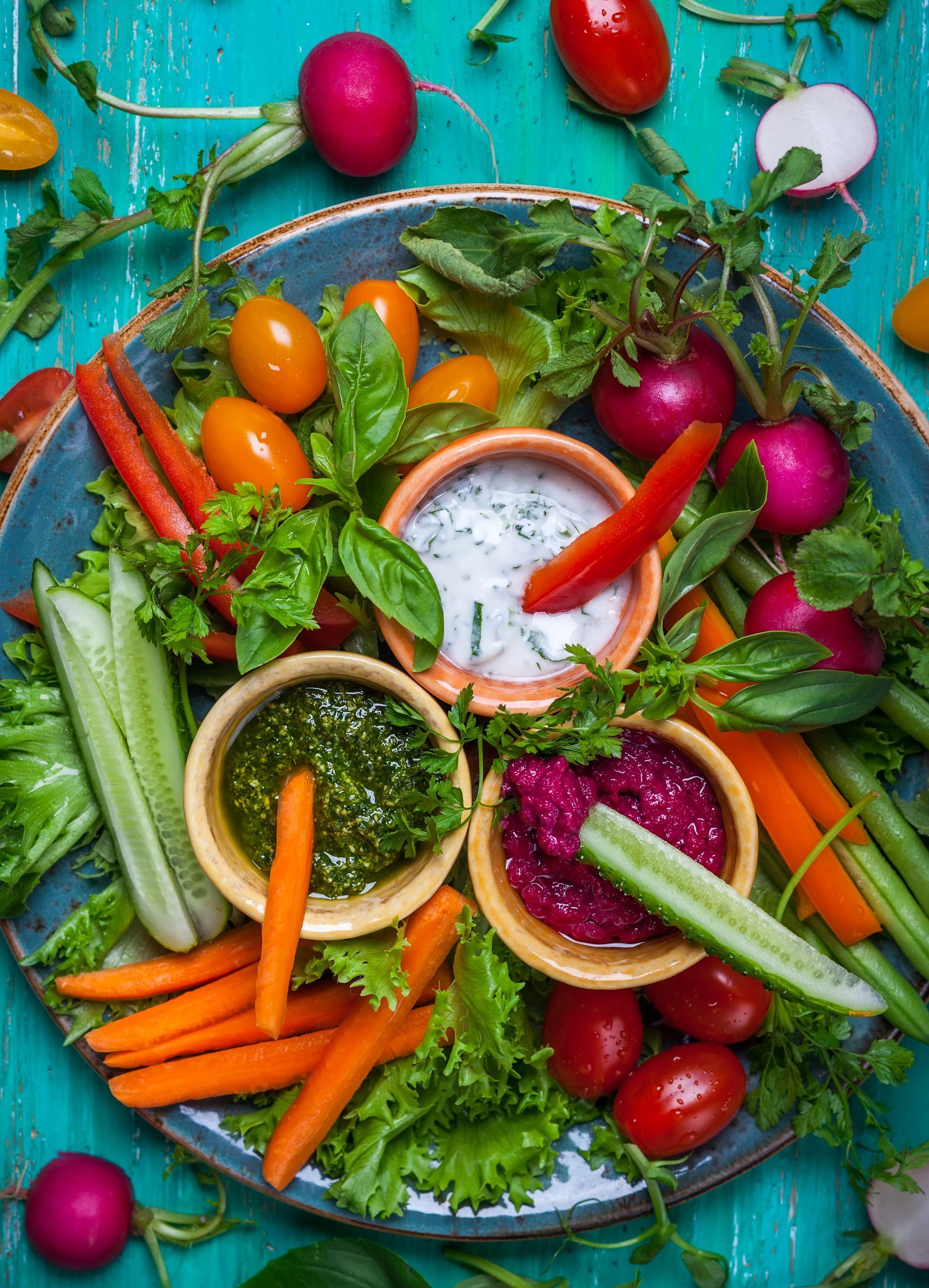 Vegetable platter with three dips: carrot sticks, cucumber slices, tomatoes, radishes, and basil leaves arranged on a blue plate.