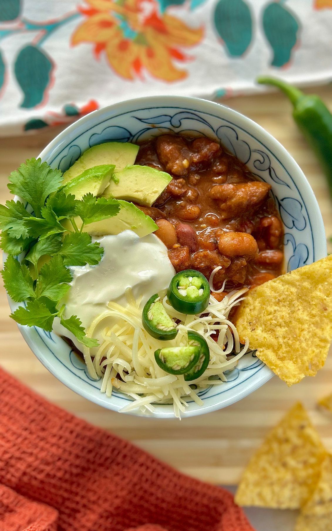 Bowl of chili topped with avocado slices, sour cream, shredded cheese, jalapeño slices, cilantro, and a tortilla chip.