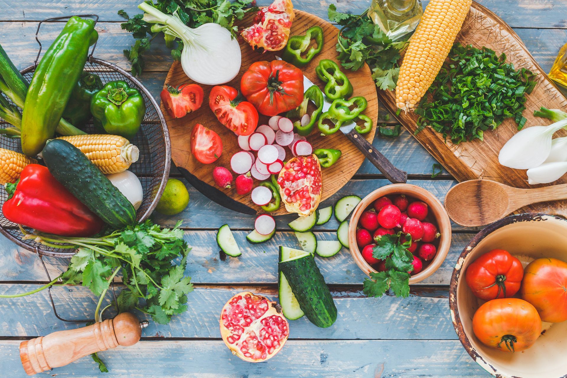 Overhead shot of colorful vegetables arranged on a blue wooden surface, including tomatoes, radishes, cucumbers, peppers, onions, and corn.