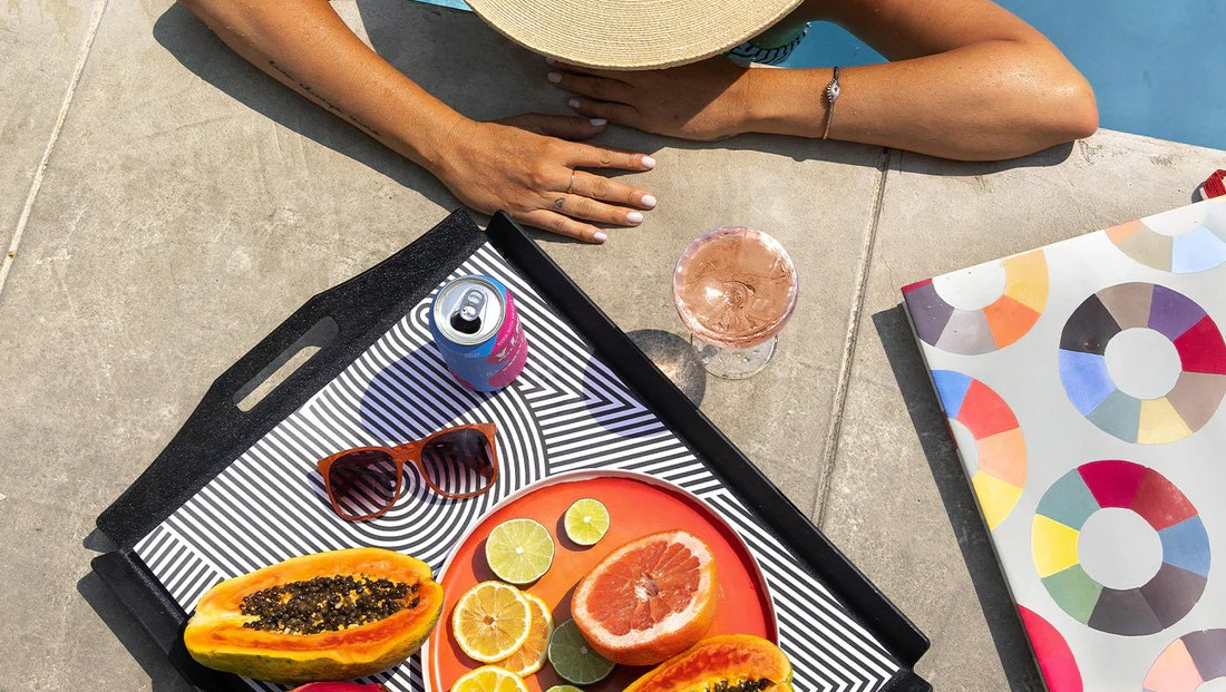 Person by a pool enjoys refreshments on a tray: fruit, drink, sunglasses, and a can, with a colorful magazine.