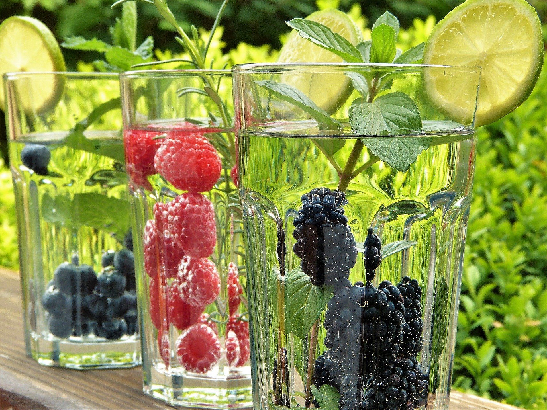 Three glasses of water infused with berries, mint, and lime, set outside with green foliage in the background.