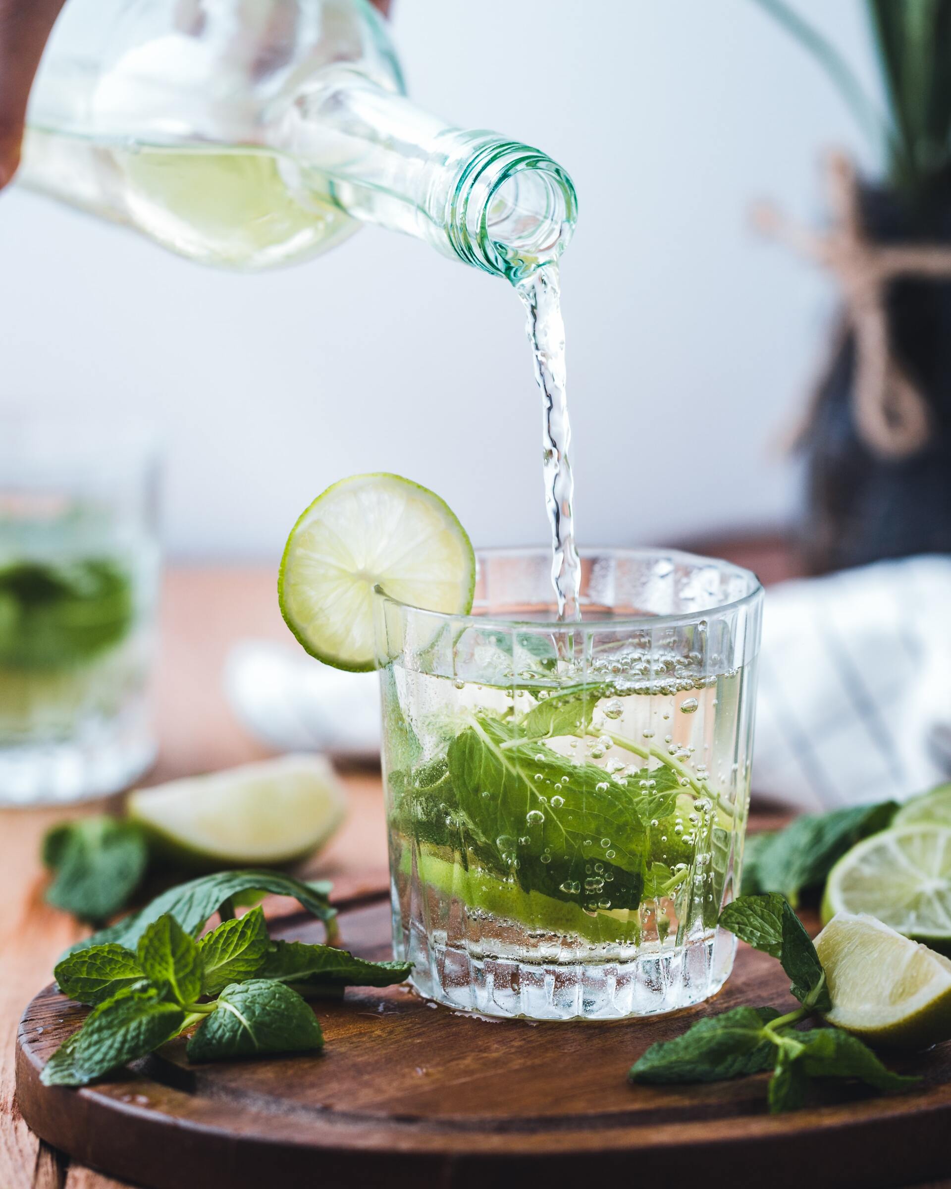 A refreshing drink being poured into a glass with mint and lime slices on a wooden tray.