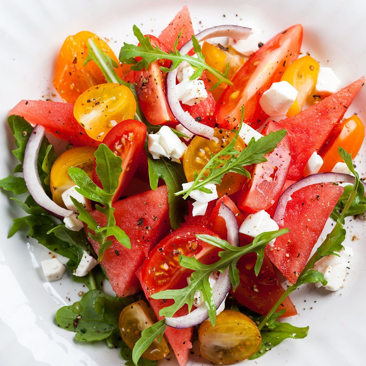 Watermelon, tomato, and feta salad with red onion and arugula in a white bowl.