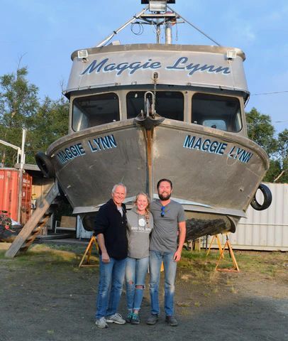 Three people stand in front of a boat named