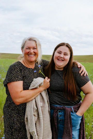 Two smiling women embracing in a field of wildflowers. One holds a flower and jacket; the other has her arm around the first.